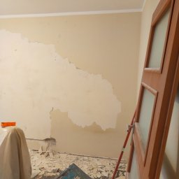 Usługi Remontowe Robert Balcerzak - Ściana w trakcie usuwania starej farby lub gładzi, widoczne fragmenty oderwanej warstwy, zakryte meble folią ochronną, skrzynka narzędziowa Makita na podłodze.