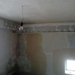 Usługi Remontowe Robert Balcerzak - Surowe wnętrze w trakcie prac wykończeniowych: ściany częściowo pokryte gładzią, widoczna żarówka na kablu, listwy przypodłogowe w trakcie montażu, małe okno w dolnej części ściany.