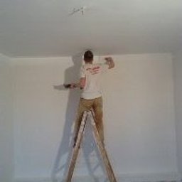 Usługi Remontowe Robert Balcerzak - Mężczyzna na drabinie maluje ścianę w pomieszczeniu. Widoczna świeżo pomalowana powierzchnia, ślady farby na podłodze zabezpieczonej folią i narzędzia malarskie.