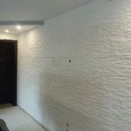 Usługi Remontowe Robert Balcerzak - Ściana wykończona białą, strukturalną okładziną przypominającą kamień, widoczne drobne niedoskonałości w połączeniach, ciemne drzwi w tle po lewej stronie, surowe oświetlenie sufitowe.