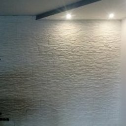 Usługi Remontowe Robert Balcerzak - Ściana pokryta dekoracyjną, białą strukturą przypominającą fale. Widoczne dwa punktowe źródła światła na suficie i fragment surowej podłogi.