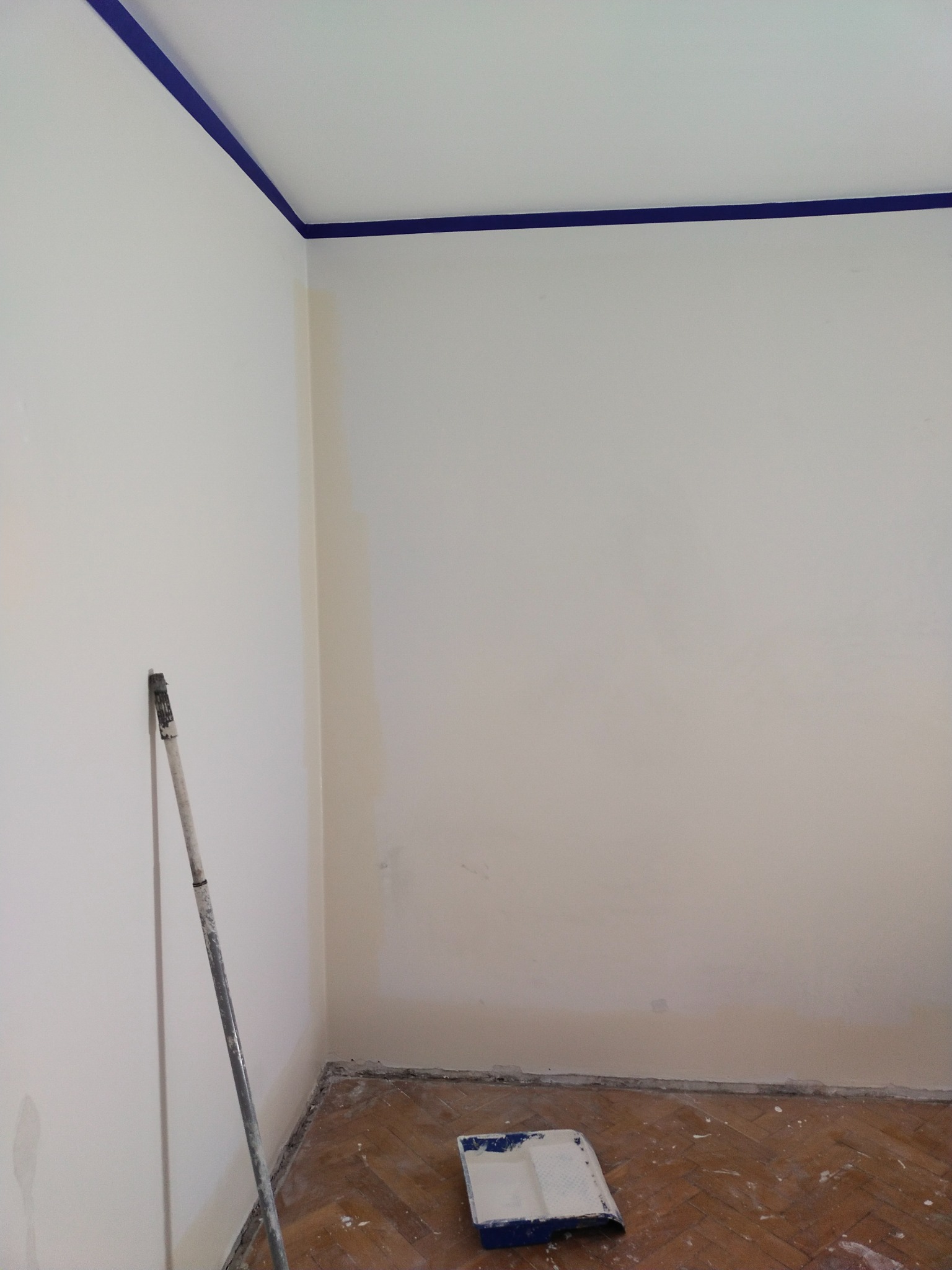 Pokój w trakcie malowania, z niebieską taśmą malarską przy suficie, wałek oparty o ścianę i kuweta z farbą na podłodze z parkietu. Ściany pomalowane na biało, widoczne ślady po starej farbie.