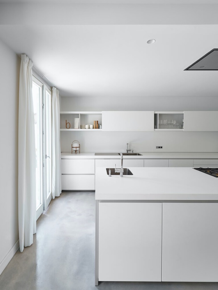 Minimalistyczna biała kuchnia z wyspą i betonową podłogą, oświetlona naturalnym światłem wpadającym przez okno z białymi zasłonami.