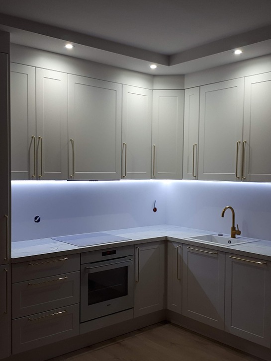 Jasnoszara kuchnia narożna z białym blatem, złotymi uchwytami i podświetleniem LED pod szafkami wiszącymi; widoczne gniazdka elektryczne w ścianie.