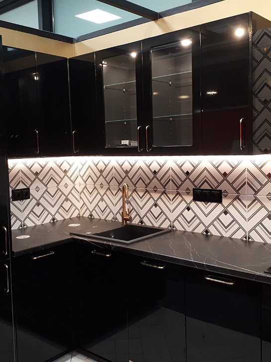 Część narożnej, czarnej zabudowy kuchennej z połyskiem, z blatem imitującym czarny marmur, złotą baterią i dekoracyjną mozaiką na ścianie.