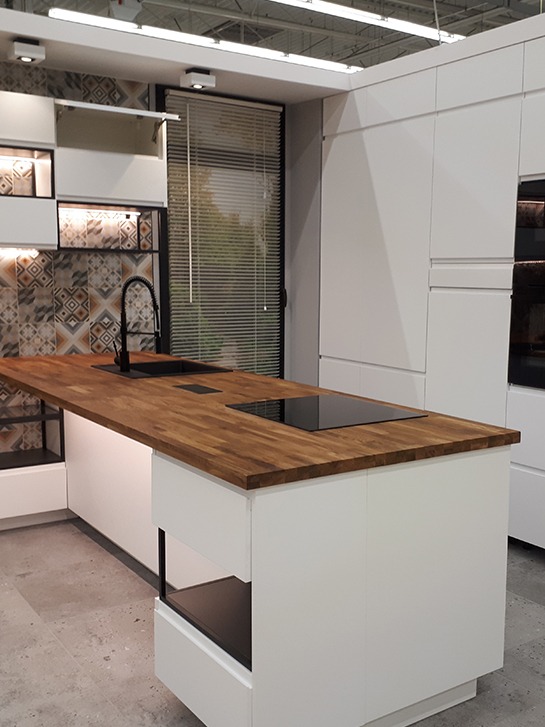 Nowoczesna kuchnia z białą zabudową i drewnianym blatem, czarny zlew i płyta indukcyjna, wzorzyste płytki nad blatem, żaluzje w oknie.