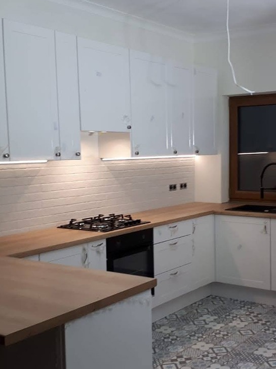 Jasna kuchnia z białymi szafkami, drewnianym blatem i wzorzystą podłogą, oświetlenie LED pod szafkami wiszącymi, czarna płyta grzewcza i piekarnik.