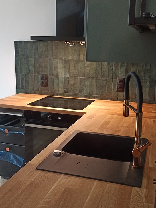 Fragment kuchni z drewnianym blatem, czarnym zlewem z baterią w kolorze miedzi, płytą indukcyjną i zielonymi płytkami na ścianie nad blatem.