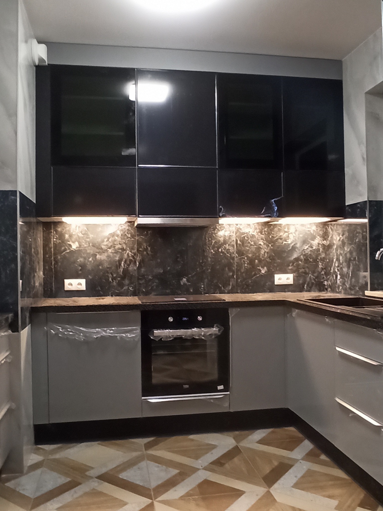 Nowoczesna kuchnia z czarnymi, błyszczącymi szafkami wiszącymi, szarymi szafkami dolnymi, blatem imitującym marmur i podświetleniem LED, częściowo jeszcze zabezpieczona folią ochronną.