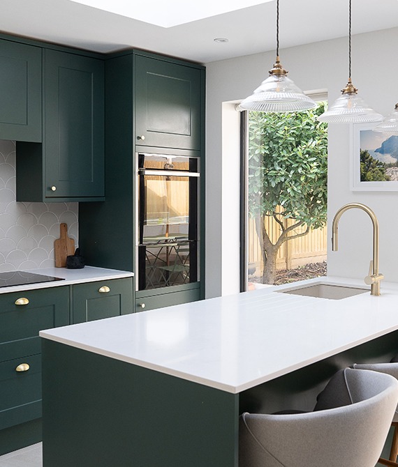 Nowoczesna kuchnia w odcieniach butelkowej zieleni z białym blatem i złotymi akcentami, widok na ogród przez duże okno, oświetlona wiszącymi lampami z ryflowanego szkła.