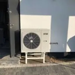 Biała pompa ciepła marki Ferroli zamontowana na białym metalowym stelażu przy ścianie budynku z otwartymi drzwiami wejściowymi po lewej stronie.