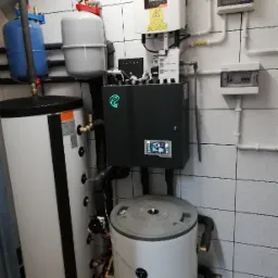 Kompletna kotłownia z dwoma zbiornikami, kotłem z wyświetlaczem i instalacją rurową w pomieszczeniu wyłożonym białymi płytkami.