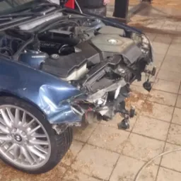 Uszkodzony przód niebieskiego BMW z otwartą maską w warsztacie, widoczne uszkodzenia karoserii i odsłonięte elementy silnika, na podłodze rozsypane drobne zanieczyszczenia.