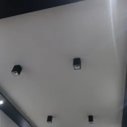 Biały sufit z czarnymi elementami dekoracyjnymi i punktowymi lampami w kwadratowych obudowach, widoczne odbicie światła.