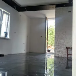 Wnętrze w trakcie prac wykończeniowych: czarna, polerowana podłoga z marmurowym wzorem, surowa ściana z cegły, ściany przygotowane do malowania, czarny pas sufitowy, otwarte drzwi na zewnątrz.