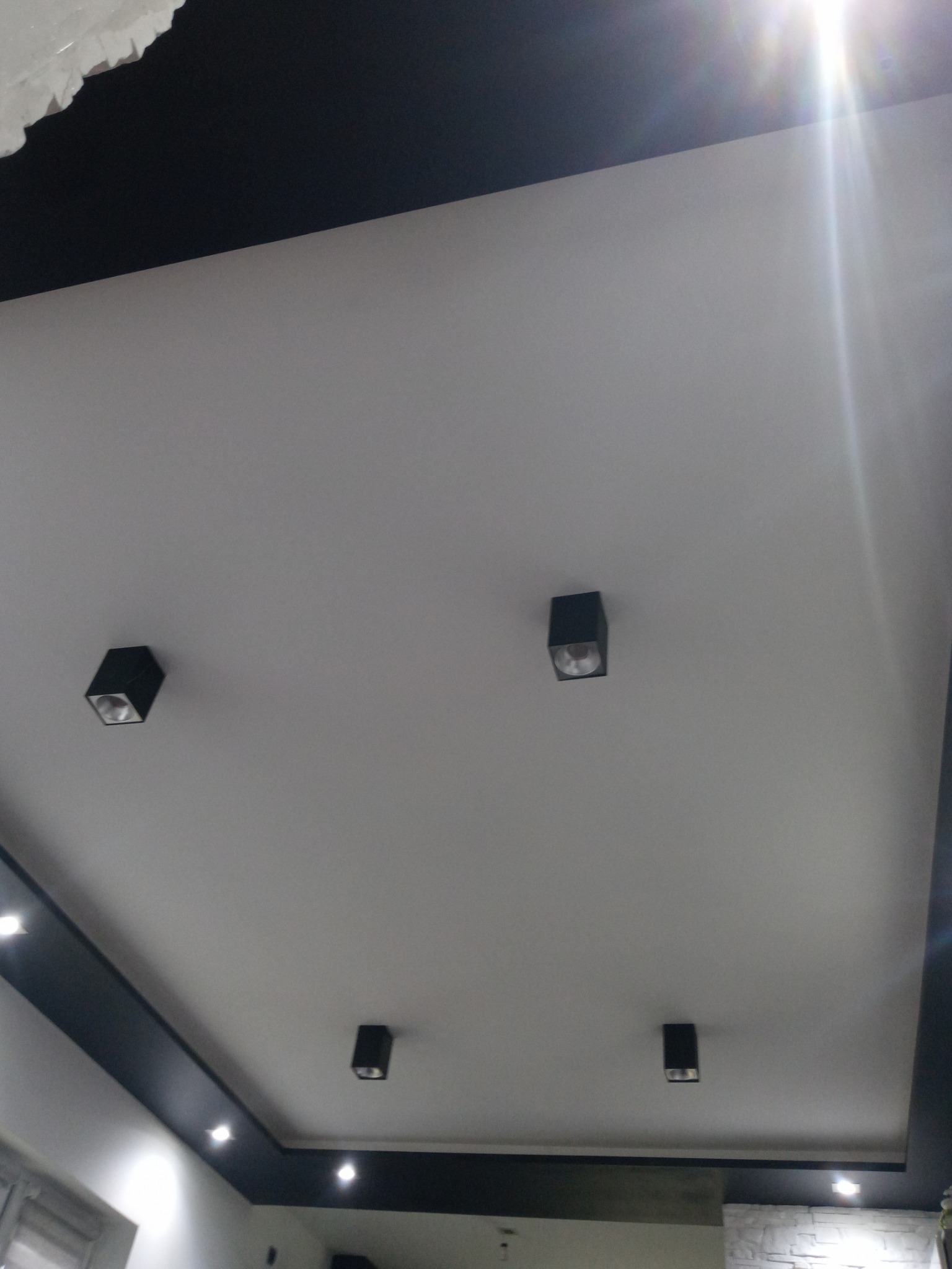 Biały sufit z czarnymi elementami dekoracyjnymi i punktowymi lampami w kwadratowych obudowach, widoczne odbicie światła.