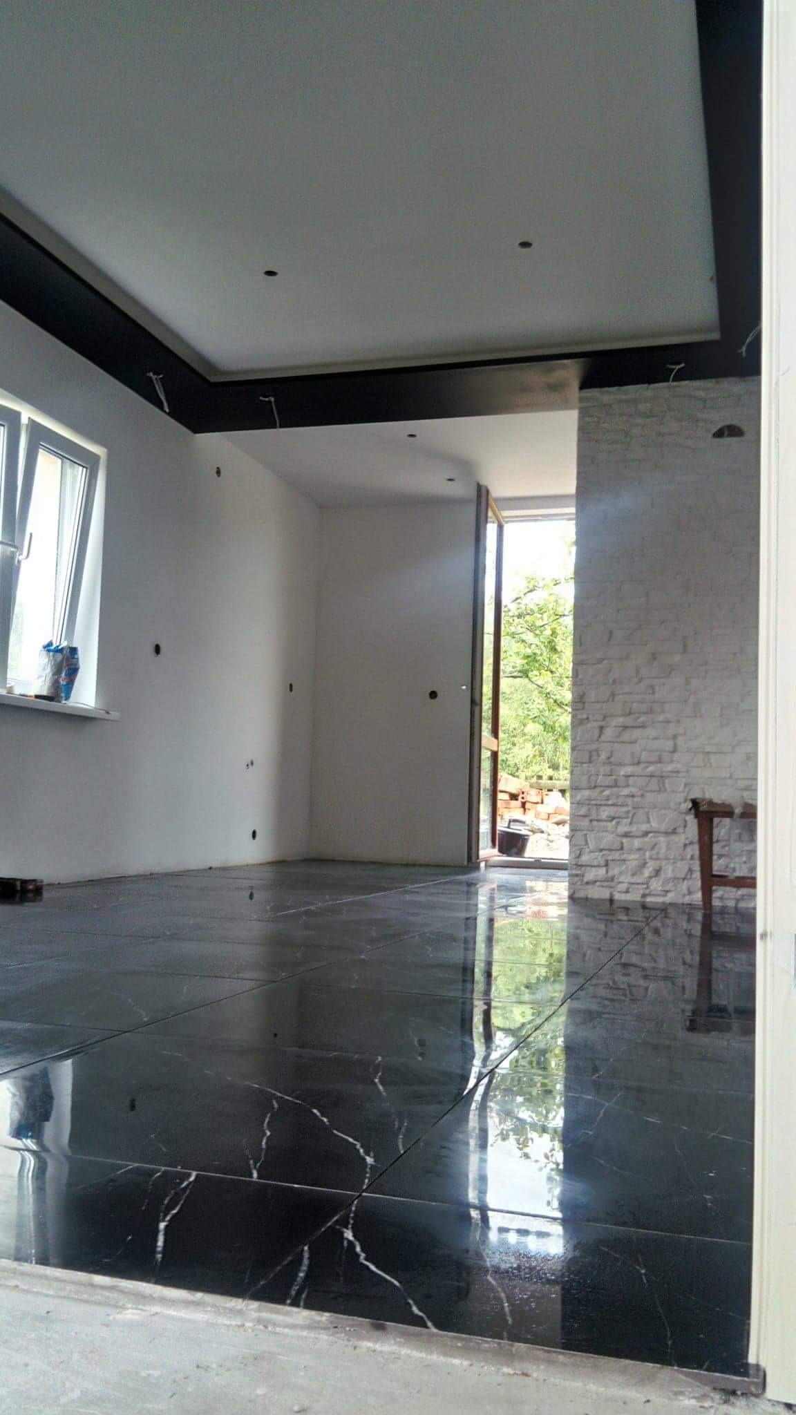 Wnętrze w trakcie prac wykończeniowych: czarna, polerowana podłoga z marmurowym wzorem, surowa ściana z cegły, ściany przygotowane do malowania, czarny pas sufitowy, otwarte drzwi na zewnątrz.