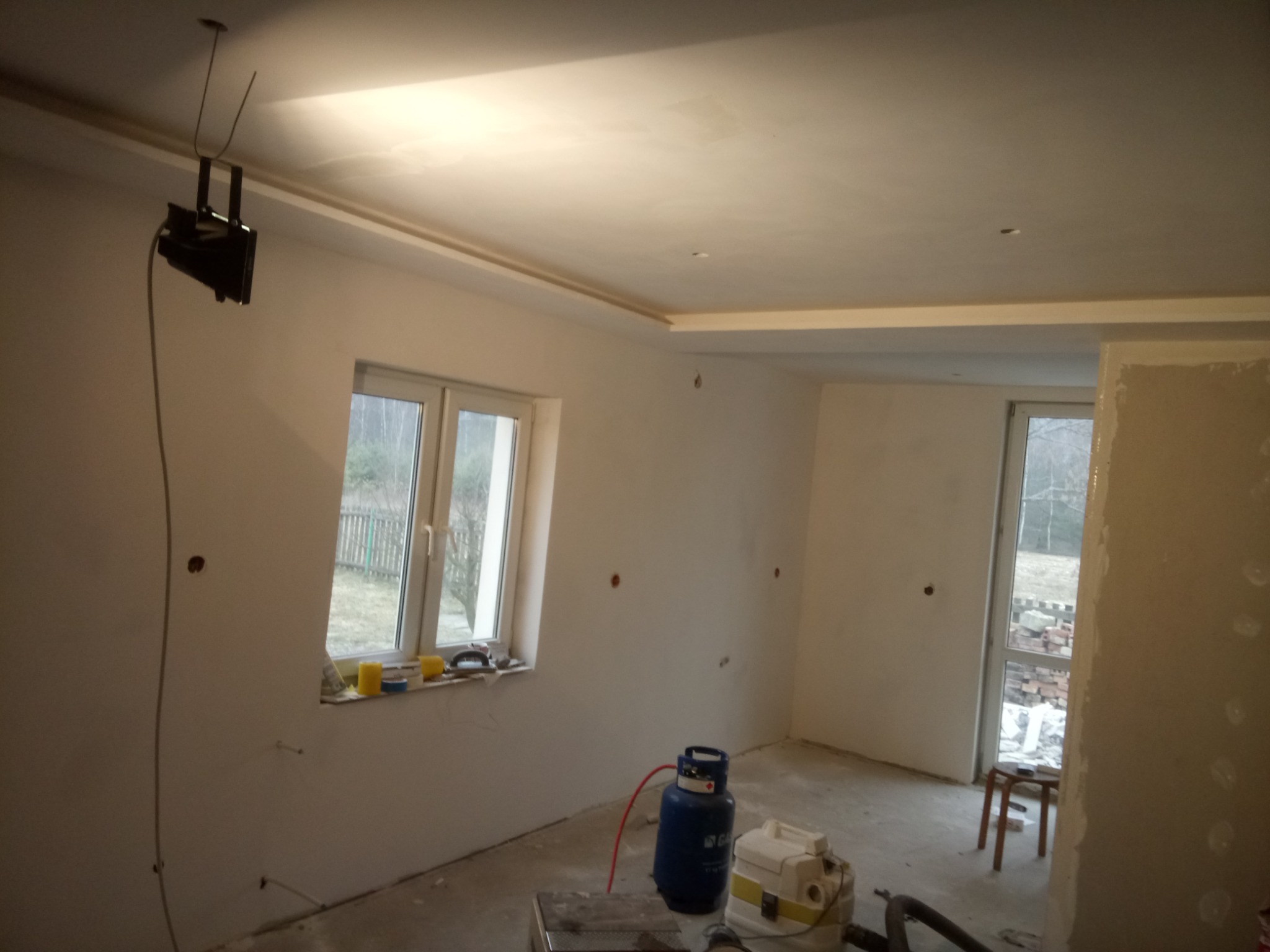 Wnętrze w trakcie prac wykończeniowych: ściany przygotowane do malowania, widoczne otwory na instalacje, listwy dekoracyjne pod sufitem, sprzęt do prac wykończeniowych, butla gazowa i prowizoryczne...