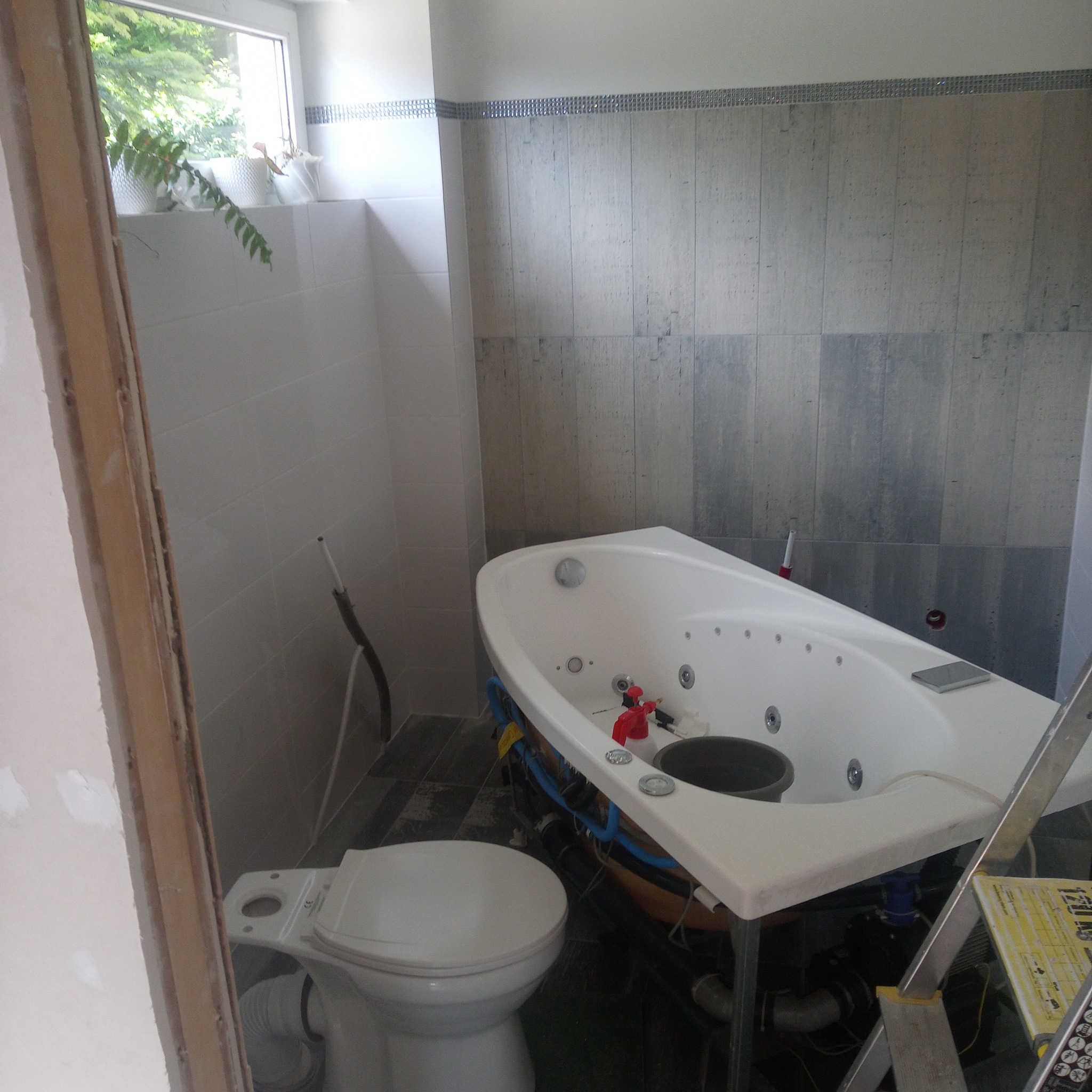 Łazienka w trakcie remontu, widoczna narożna wanna z hydromasażem na podwyższeniu, częściowo podłączona instalacja, obok biała toaleta, w tle szare płytki imitujące drewno i okno z roślinami.