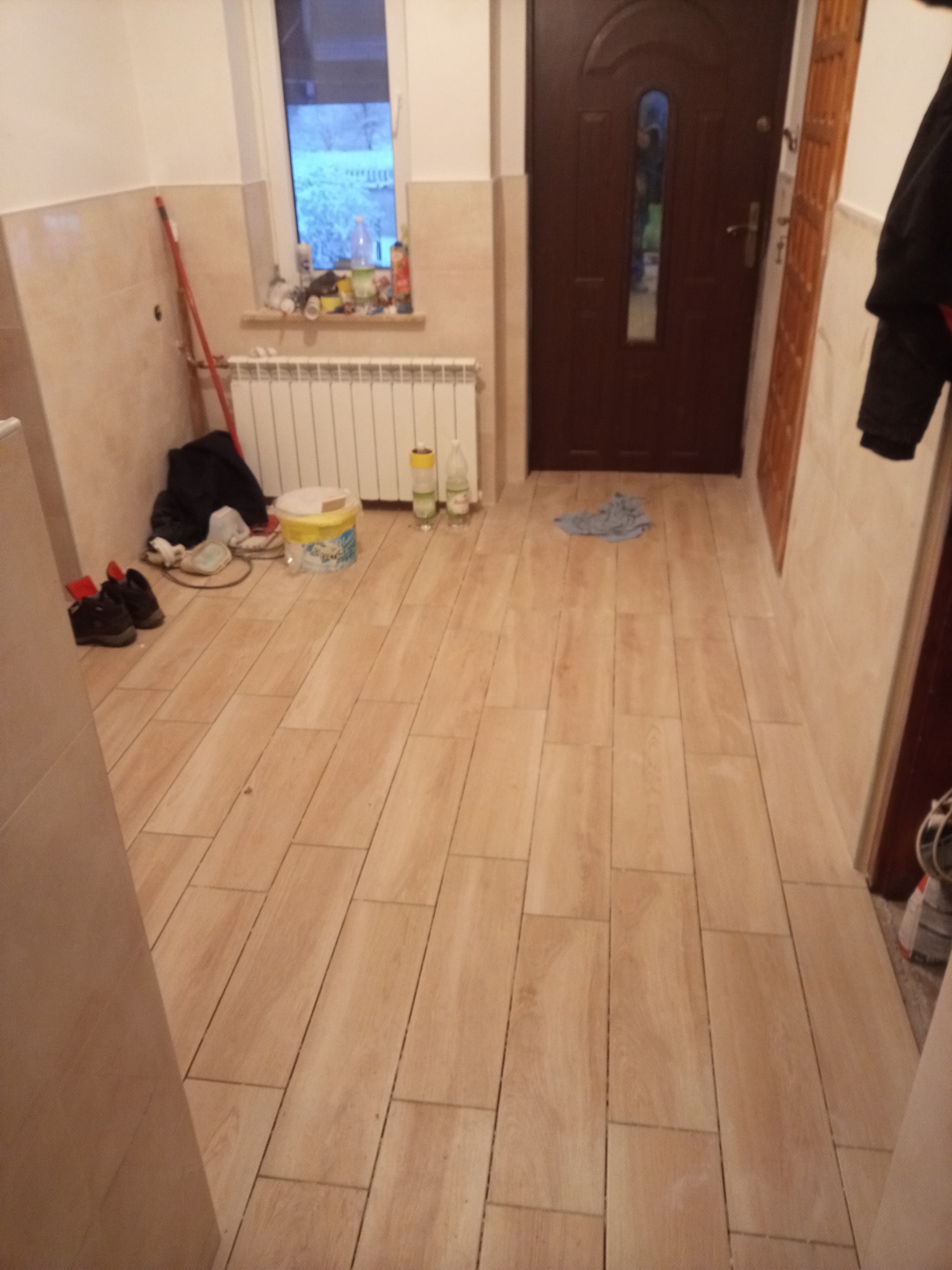 Ułożone podłużne płytki podłogowe imitujące drewno w przedpokoju, widoczne drzwi wejściowe, grzejnik, okno, oraz akcesoria malarskie i buty.
