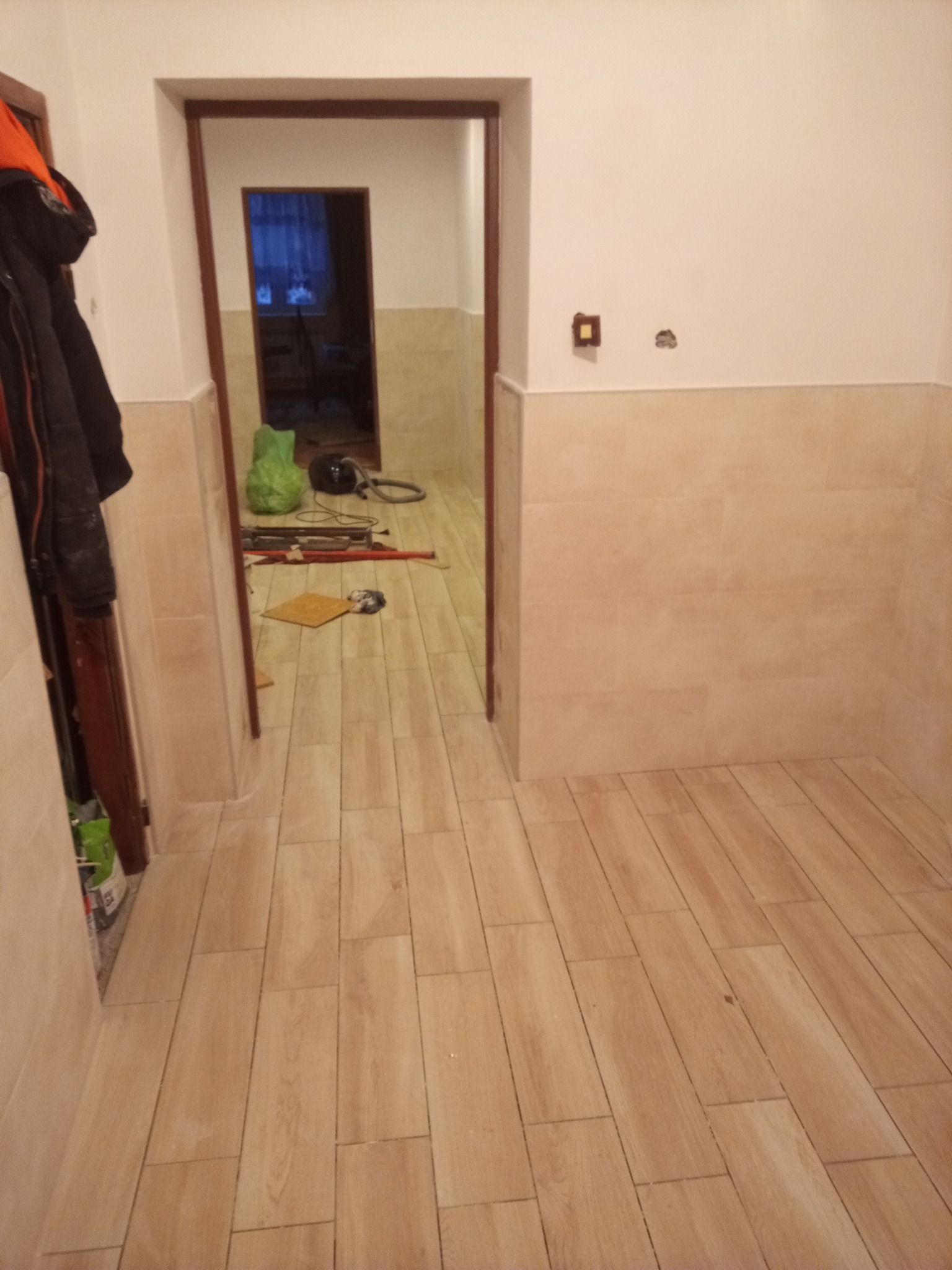 Wykończone wnętrze łazienki z beżowymi płytkami imitującymi drewno na podłodze i części ścian, widoczne narzędzia i sprzęt w dalszej części pomieszczenia.