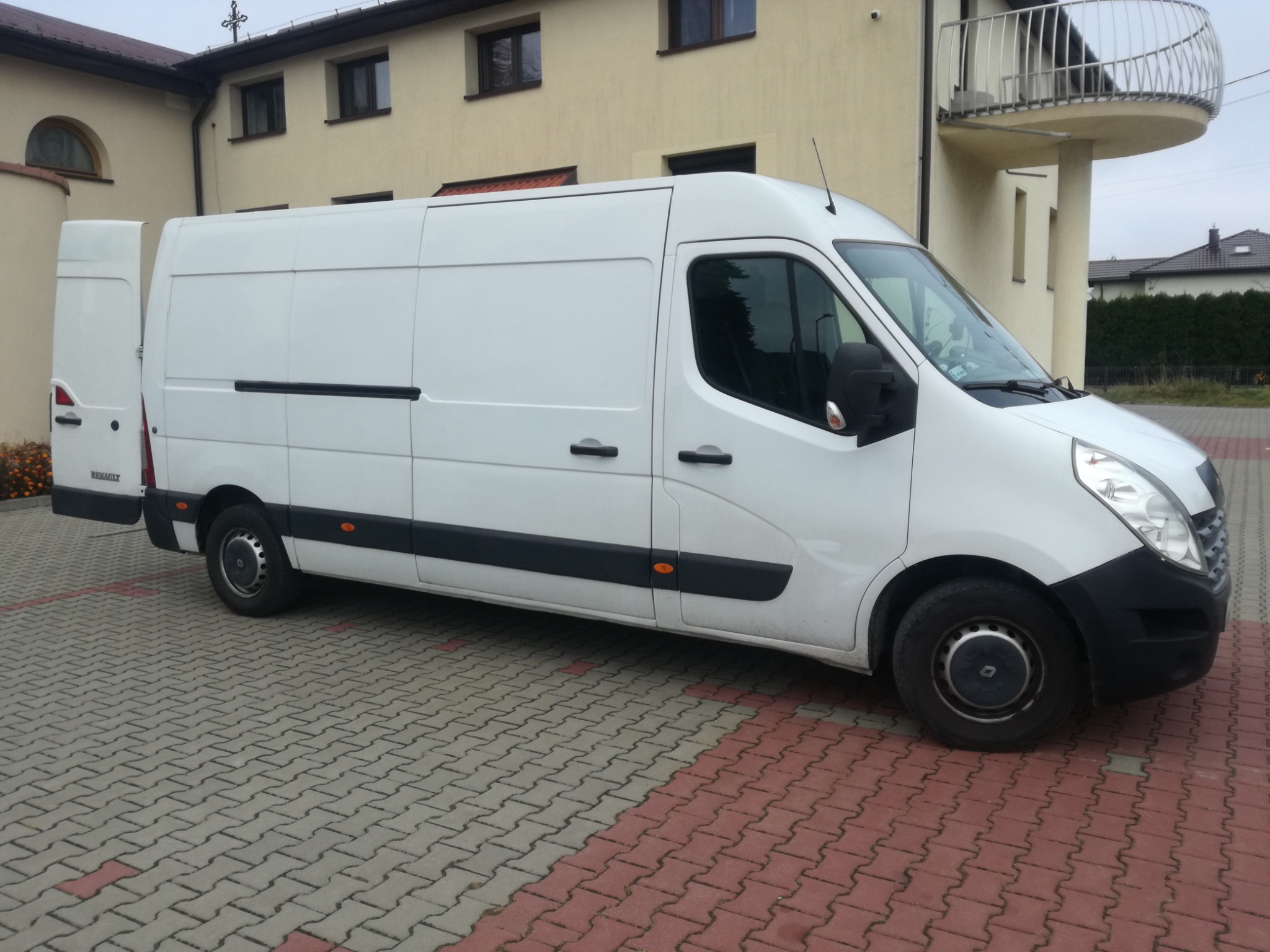 Biały samochód dostawczy Renault Master z otwartymi tylnymi drzwiami, zaparkowany na kostce brukowej w odcieniach szarości i czerwieni, z budynkiem w tle.