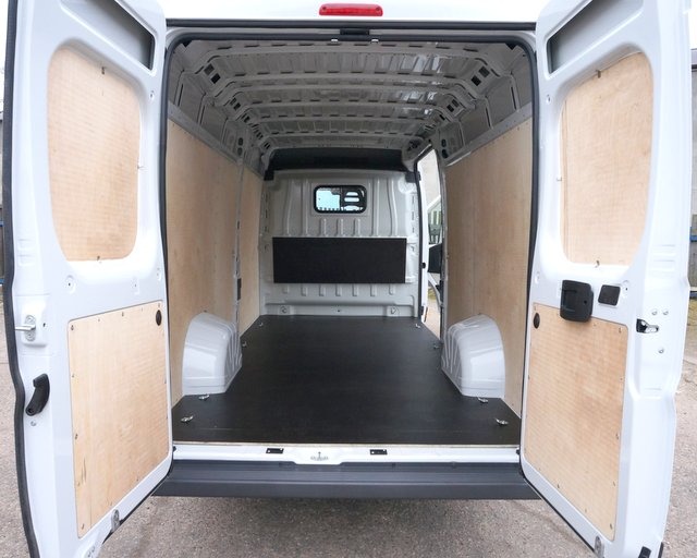 Wnętrze białego furgonetki z otwartymi tylnymi drzwiami, wyłożone sklejką na ścianach i czarną podłogą, widok od tyłu pojazdu.