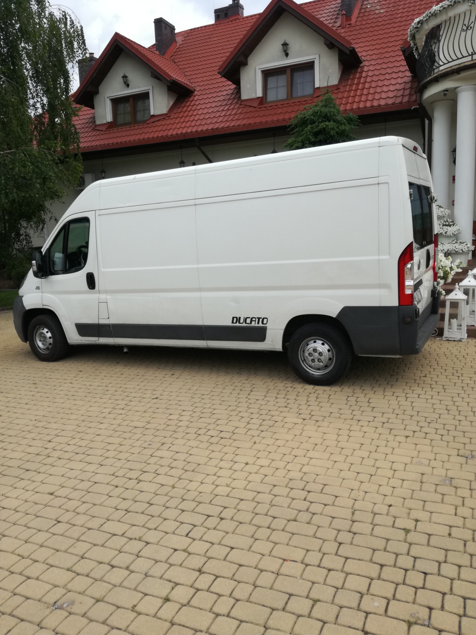 Biały furgon Ducato zaparkowany na kostce brukowej przed budynkiem z czerwoną dachówką i kolumnami.