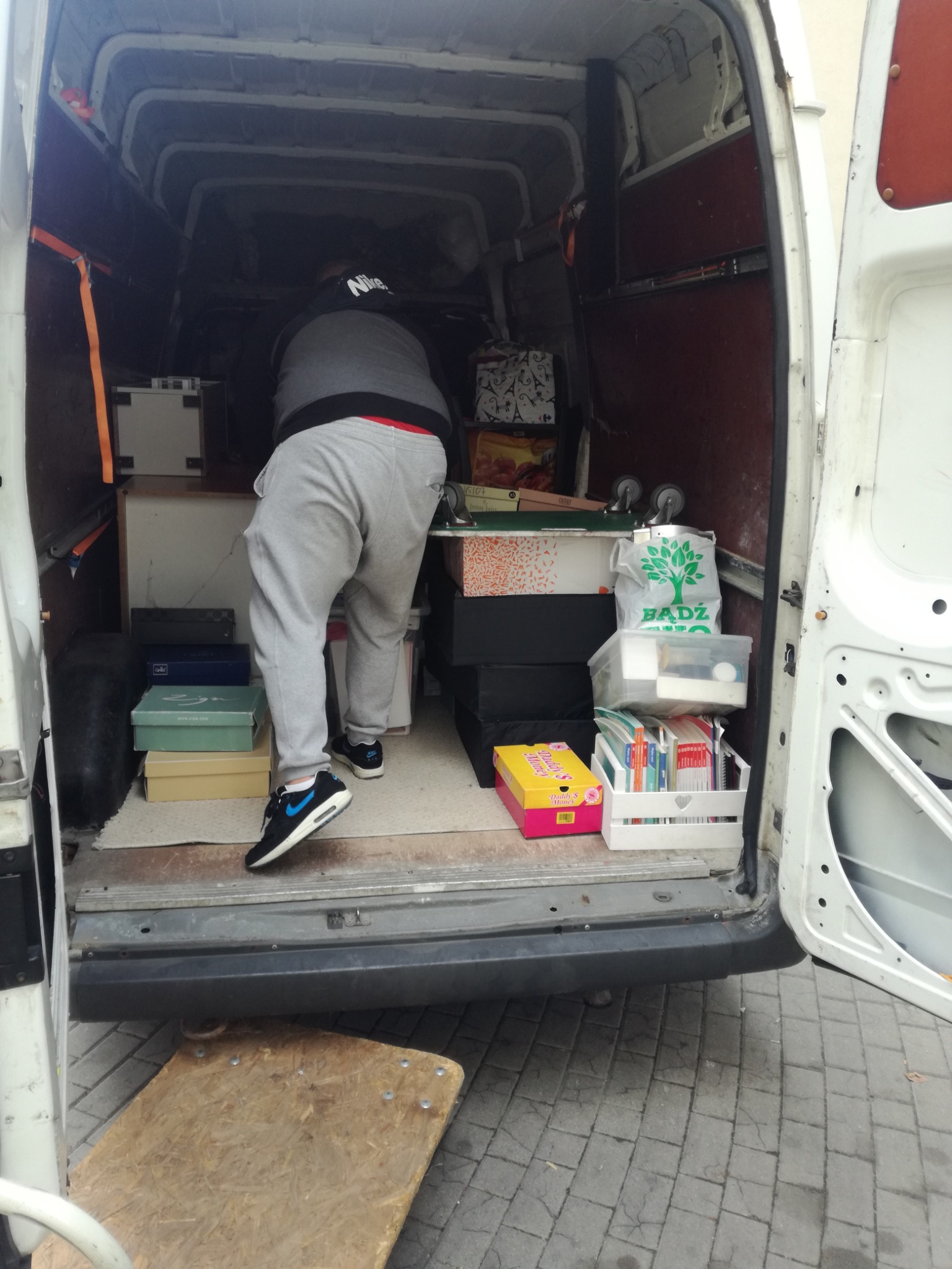 Osoba w sportowych butach wchodzi do białego busa wypełnionego kartonami i pudełkami, przygotowanego do transportu.