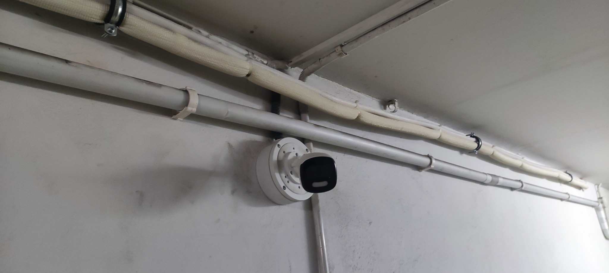 Kamera monitoringu przymocowana do ściany pod rurami instalacyjnymi, widok z dołu, szara ściana w tle.