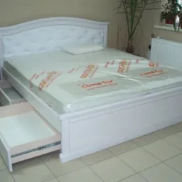 Białe łóżko z szufladami, materac w folii ochronnej, tapicerowane wezgłowie z guzikami, częściowo wysunięta szuflada, widoczne prowadnice szuflady.