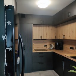 Gortex - Nowoczesna kuchnia z ciemnymi szafkami dolnymi i drewnopodobnymi górnymi, oświetlona lampą sufitową. Na blacie widoczny czajnik elektryczny i ekspres do kawy.