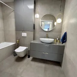 Nowoczesna łazienka w odcieniach szarości z wiszącą toaletą, szafką z umywalką nablatową, okrągłym lustrem i złotymi akcentami w postaci armatury i lamp.