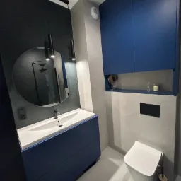 Nowoczesna łazienka w odcieniach szarości i granatu: wisząca szafka z blatem i umywalką, okrągłe lustro z podświetleniem, toaleta podwieszana, minimalistyczne oświetlenie.