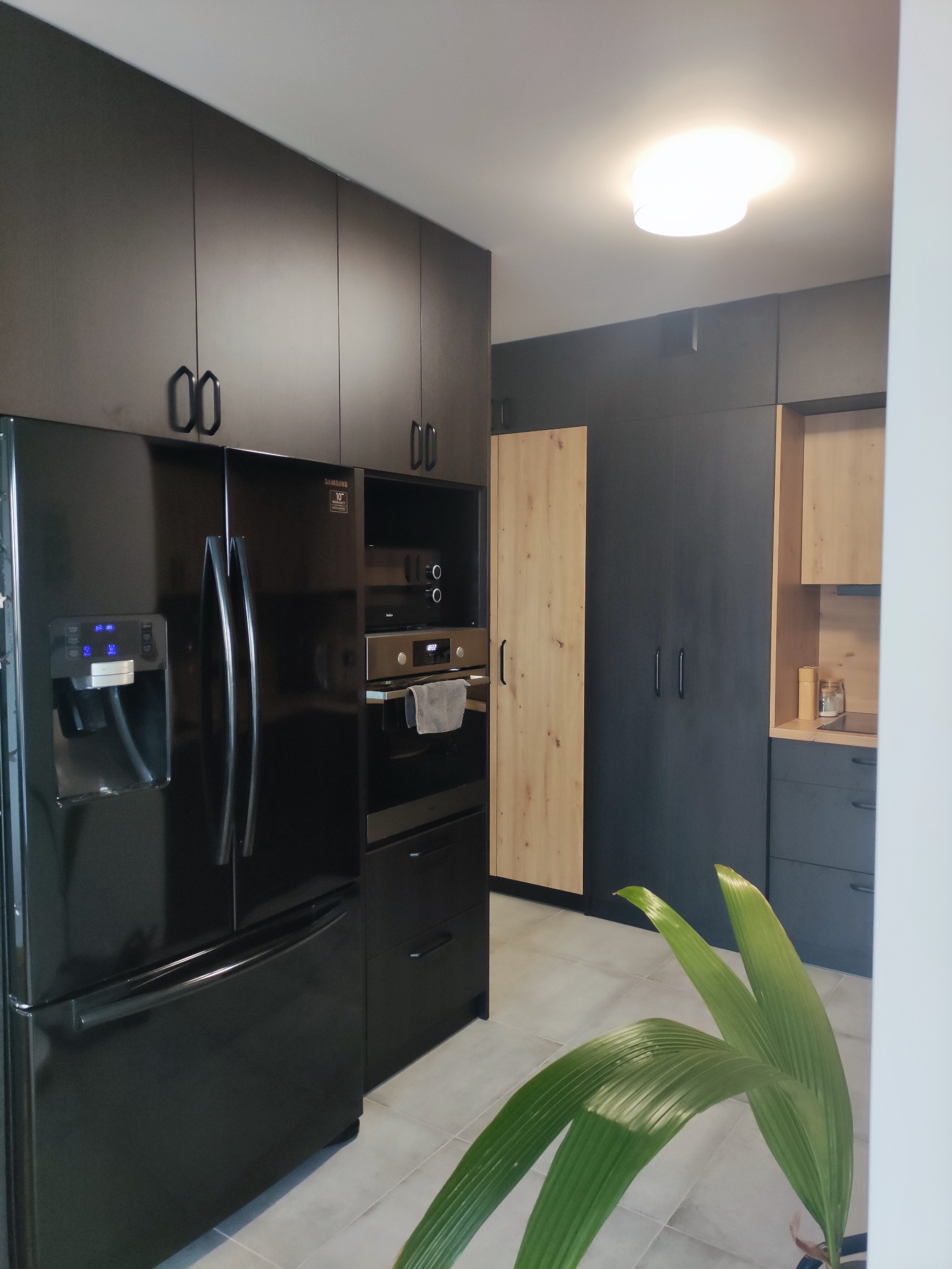 Nowoczesna kuchnia z czarnymi frontami szafek, zabudowaną lodówką Samsung z dystrybutorem wody i piekarnikiem z mikrofalówką, uzupełniona elementami z jasnego drewna i rośliną doniczkową.