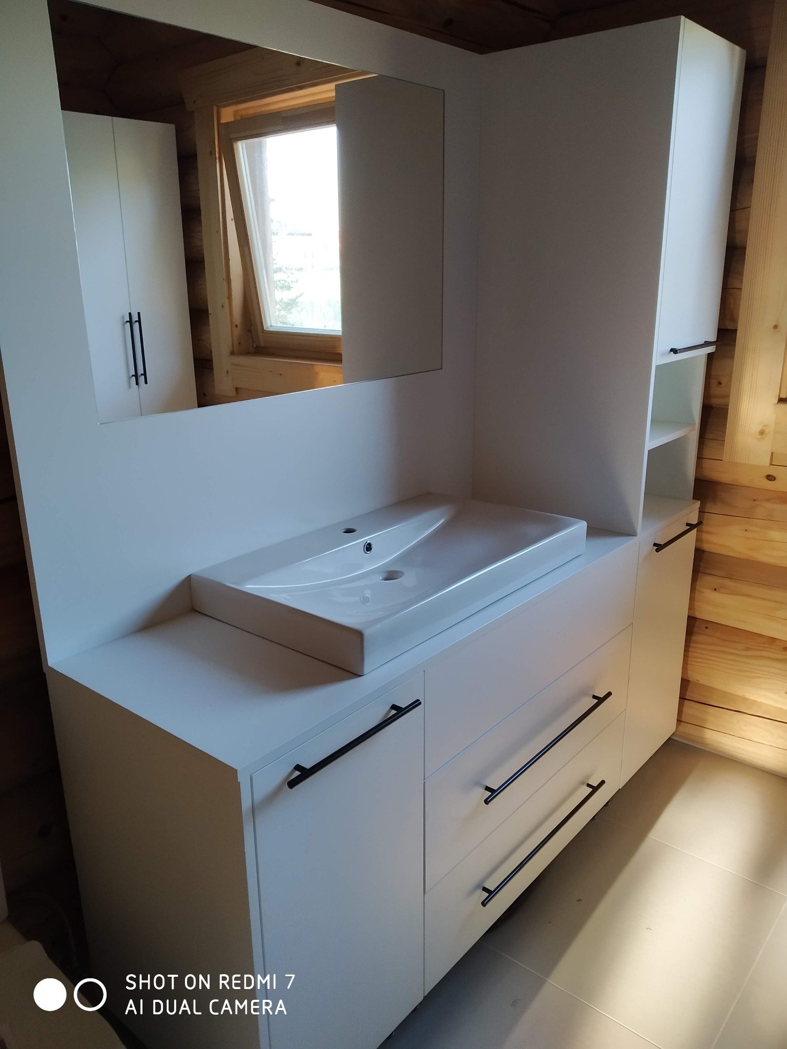 Biała, nowoczesna szafka łazienkowa z umywalką nablatową i czarnymi uchwytami, zabudowana w drewnianym wnętrzu z oknem.