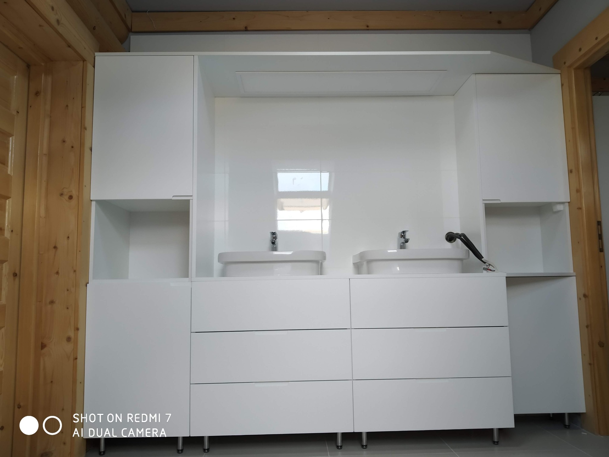 Biała szafka łazienkowa z dwiema umywalkami, trzema rzędami szuflad i górnymi szafkami, na tle białych płytek i drewnianej framugi drzwi.