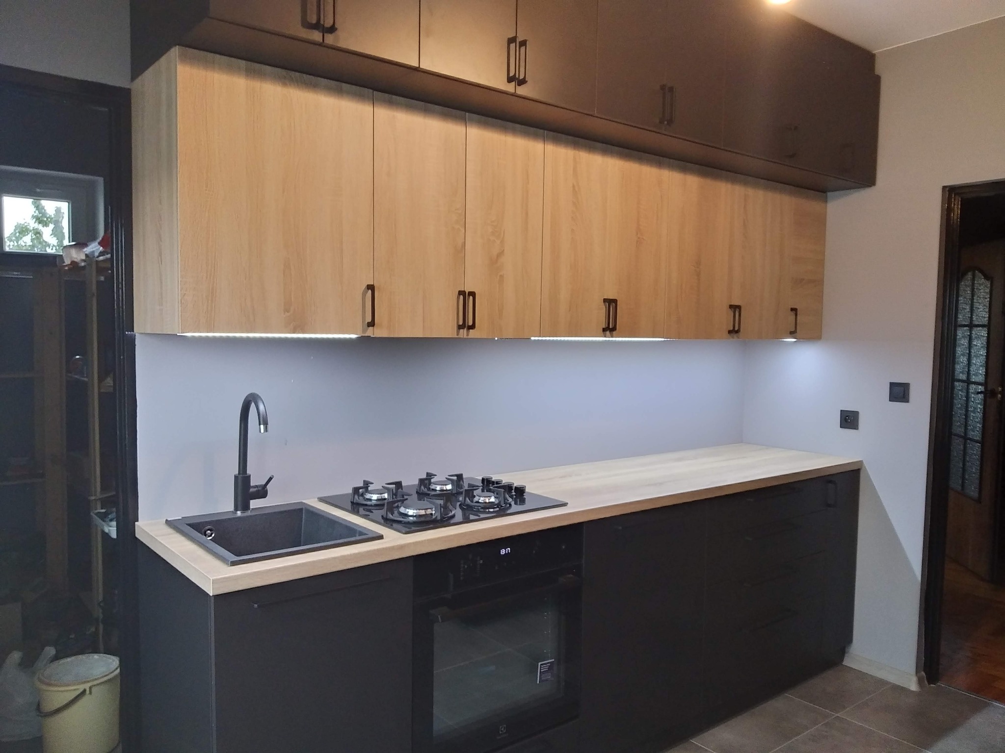 Nowoczesna kuchnia z dwukolorowymi szafkami, jasnym blatem, czarnym zlewem i płytą grzewczą oraz oświetleniem LED pod szafkami wiszącymi. Widoczny piekarnik w zabudowie.