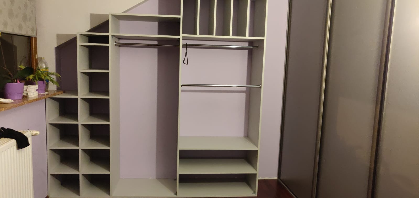Szara garderoba z otwartymi półkami i drążkami na ubrania, zintegrowana z szafą przesuwną, na tle fioletowej ściany.