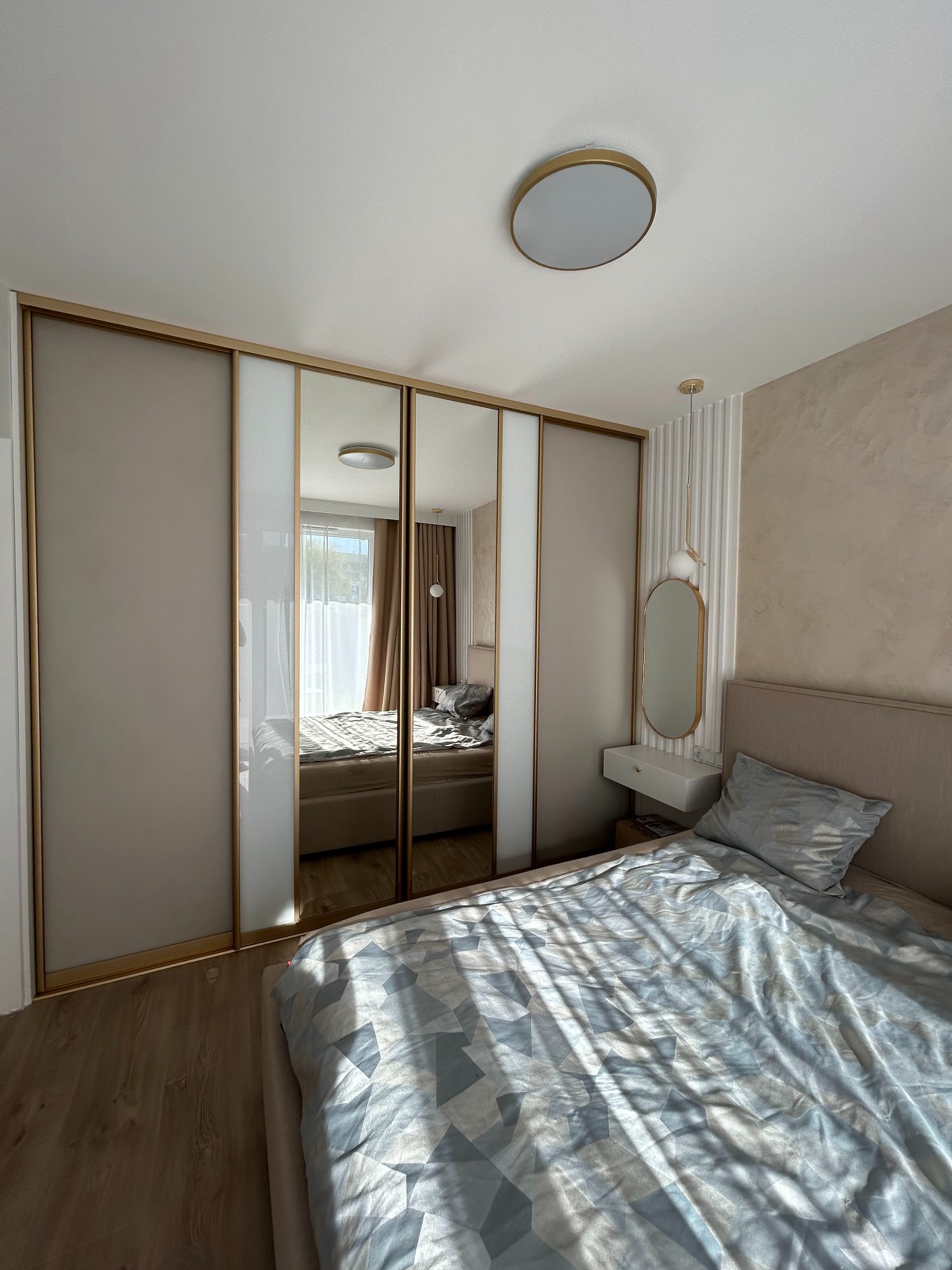 Sypialnia z beżową szafą wnękową z lustrzanymi drzwiami przesuwnymi i złotymi ramami, odbijająca łóżko z geometryczną pościelą.