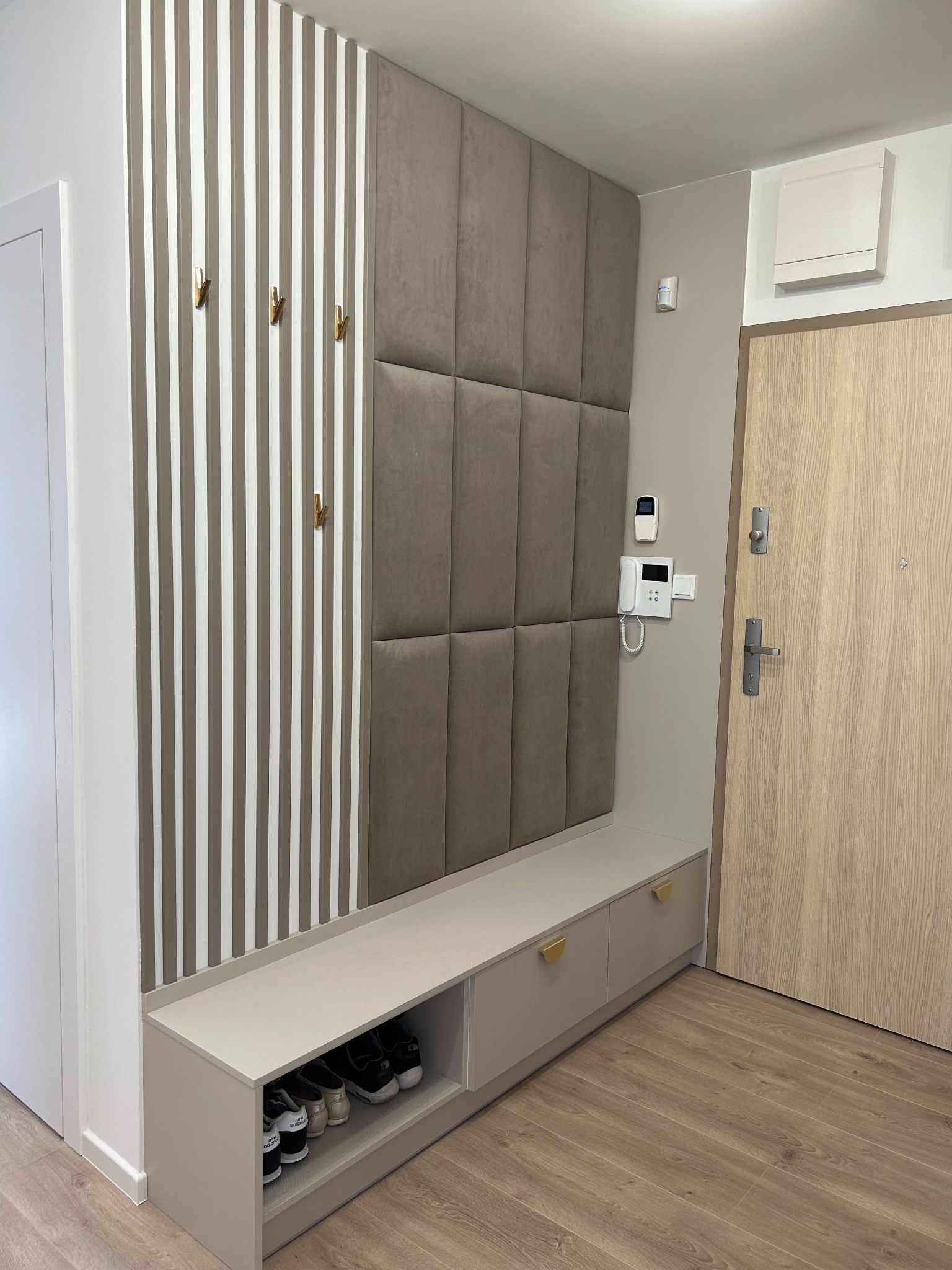 Nowoczesna aranżacja przedpokoju z siedziskiem, szafkami z półokrągłymi uchwytami, panelem tapicerowanym w kwadraty, ścianą z pionowych listew z wieszakami i drzwiami wejściowymi.