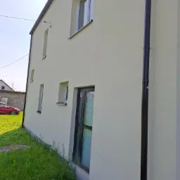 Jasna elewacja domu jednorodzinnego z brązową rynną, widok z boku na trawnik i częściowo widoczny czerwony samochód w tle. Minimalistyczna architektura.