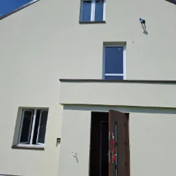 Elewacja domu jednorodzinnego w Stopnicy, z otwartymi drzwiami wejściowymi i oknami. Jasny tynk, ciemne obramowania dachu i okien.