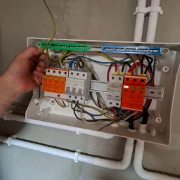 Otwarta skrzynka elektryczna z zamontowanymi bezpiecznikami i przewodami, instalowana na szarej ścianie. Widoczne białe rury instalacyjne.