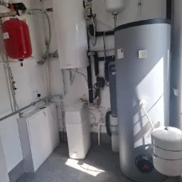 Zestaw instalacji grzewczej w kotłowni: zbiorniki, rury, filtry. Widoczne elementy instalacji CO, zasilania i filtry wody. Rzut z dołu na szarą podłogę.