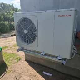 Biała pompa ciepła Immergas zamontowana na metalowej konstrukcji przy ścianie budynku z numerem 49, widok z boku, częściowo widoczna osoba podczas montażu.