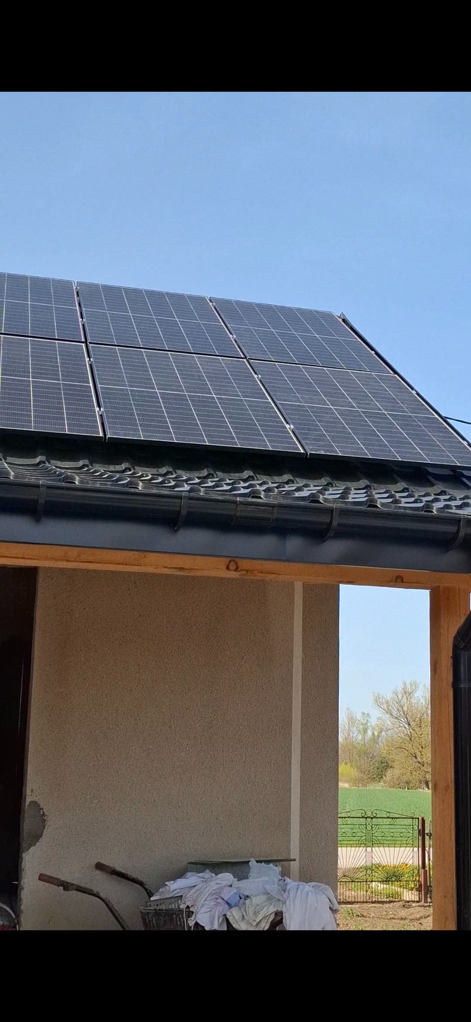 Panele słoneczne na dachu budynku z beżową elewacją. W tle błękitne niebo i zielone pole. Częściowo widoczny wózek budowlany.