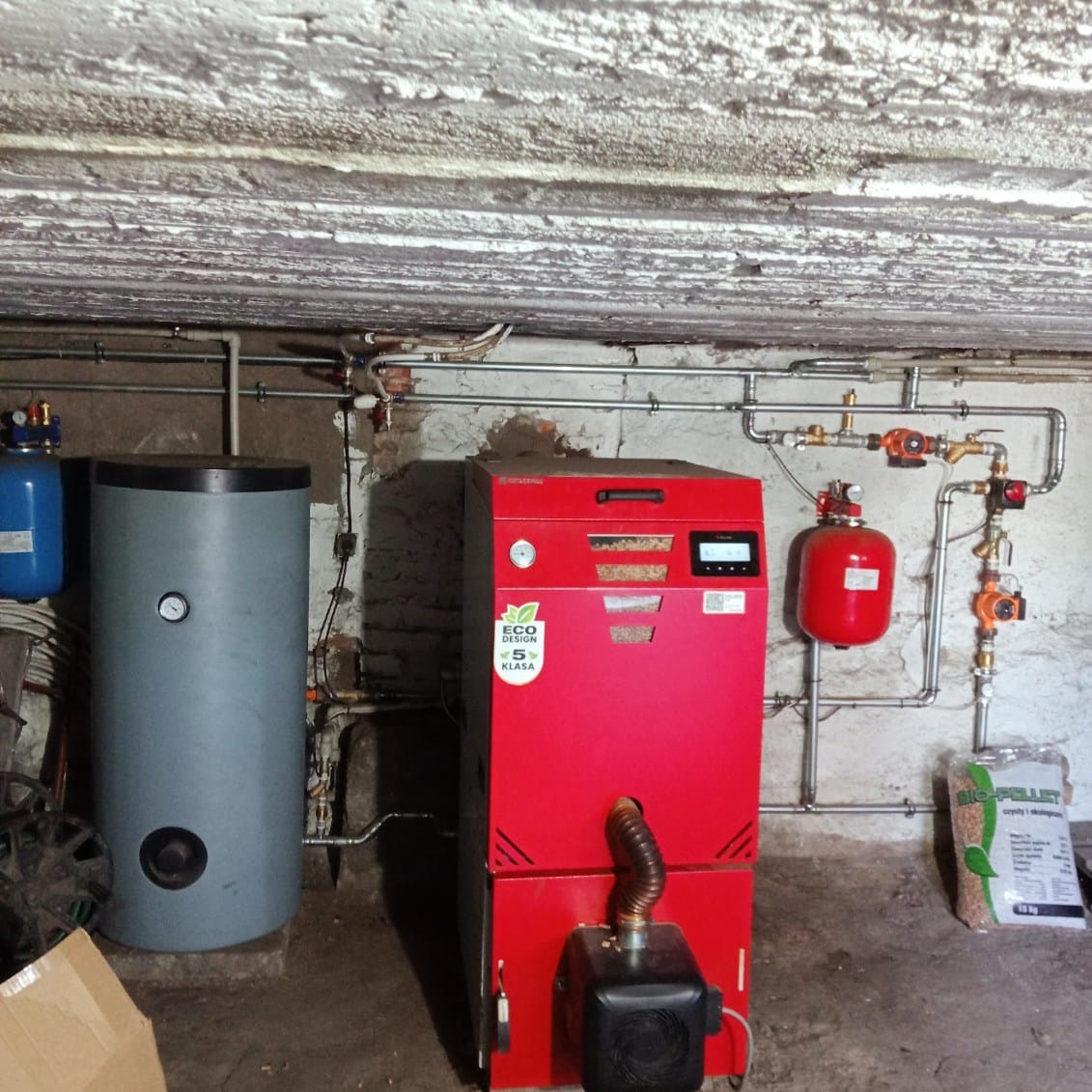 Czerwony piec na pellet z podajnikiem, szary zbiornik akumulacyjny i instalacja CO w piwnicy. Widoczny worek z pelletem.