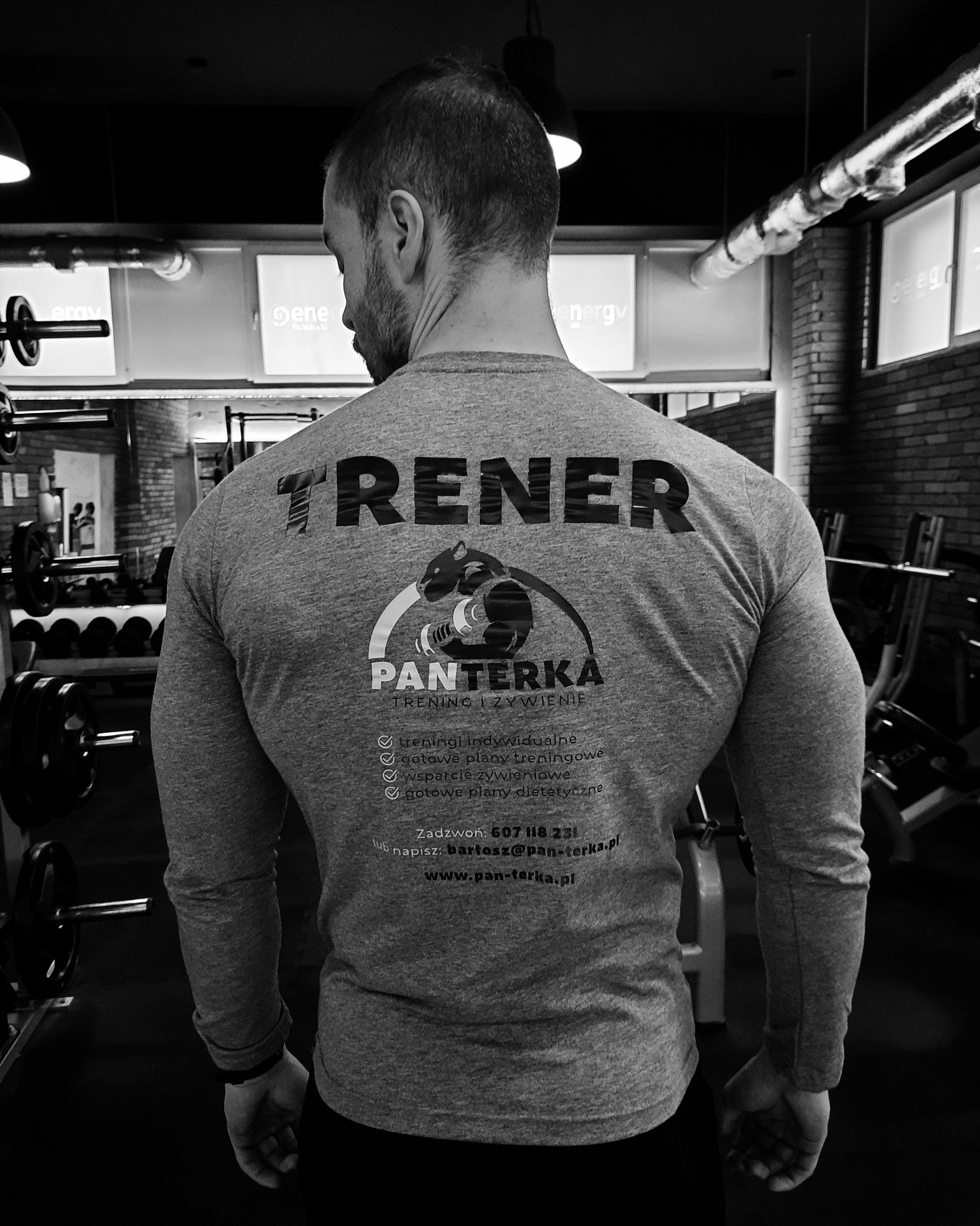 Mężczyzna o muskularnej sylwetce, odwrócony tyłem, w szarej koszulce z napisem 'TRENER' i logo firmy 'PANTERKA trening i żywienie' oferującej indywidualne treningi, plany treningowe, wsparcie...