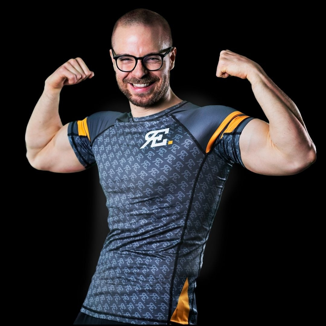 Uśmiechnięty mężczyzna w okularach, napinający bicepsy, ubrany w sportową koszulkę z wzorem i logo na czarnym tle.